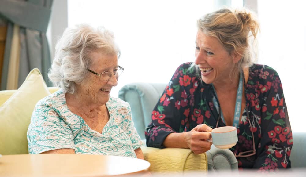 A female staff member and an elderly woman are seated in a chair, enjoying each other's company and engaging in conversation.