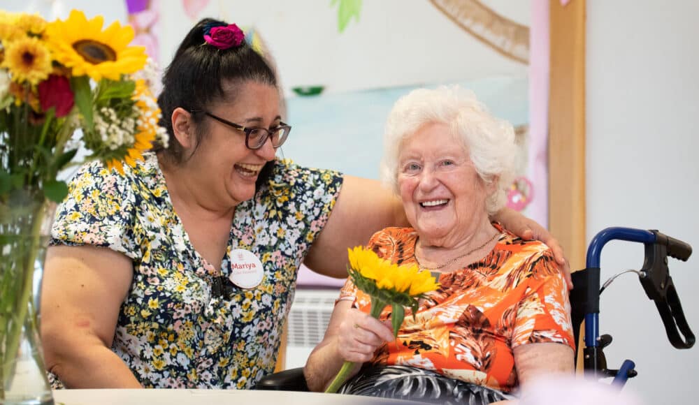 A smiling female resident holds a flower beside a female staff member, sharing a joyful moment together.