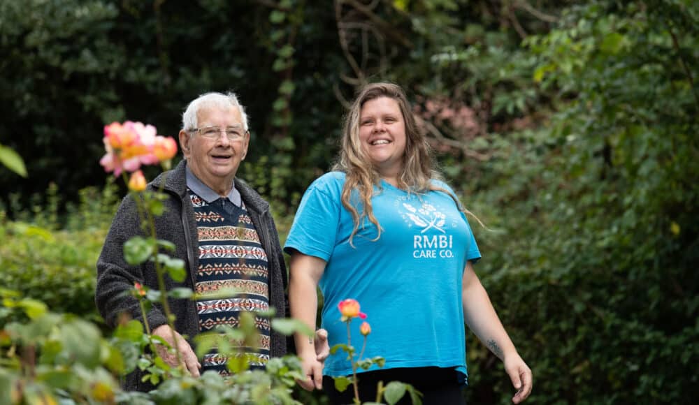 A make resident and female staff member stroll together through a vibrant garden filled with blooming flowers and lush greenery.