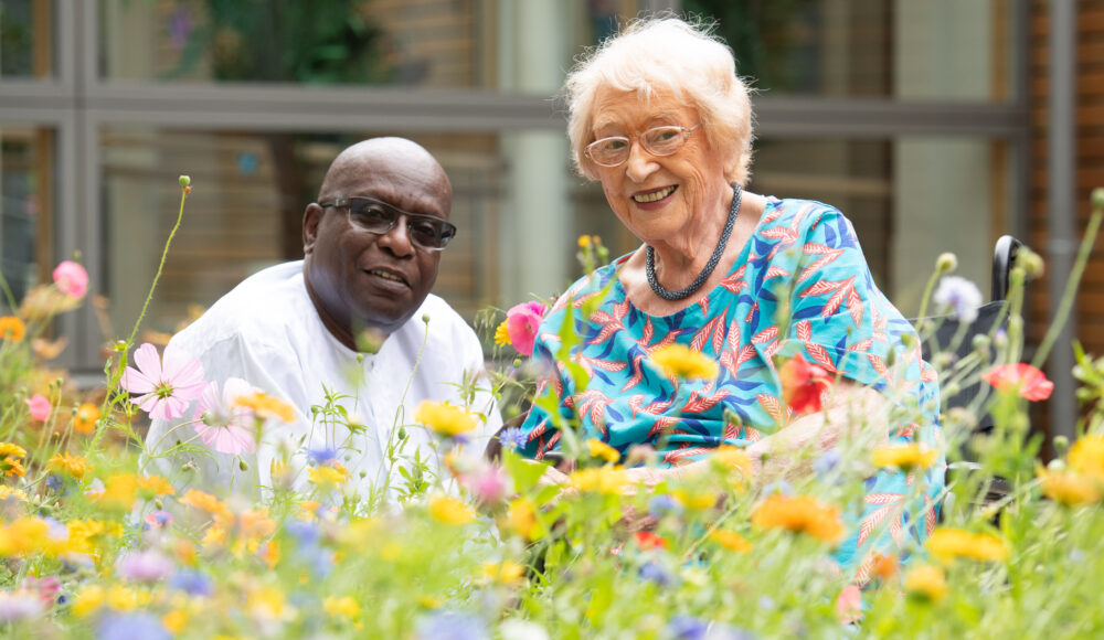 A staff member and a resident sits in a flower-filled garden, surrounded by benches and a gazebo, enjoying the tranquility of nature.