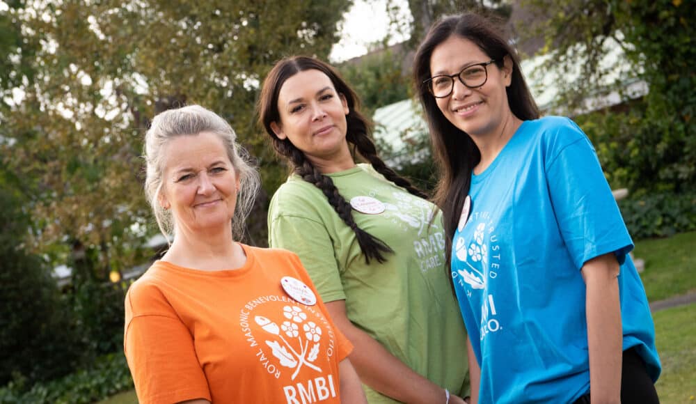 Three female staff members stand together, wearing bright orange, blue, and green shirts, smiling and enjoying each other's company at Prince George Duke of Kent Court care home.