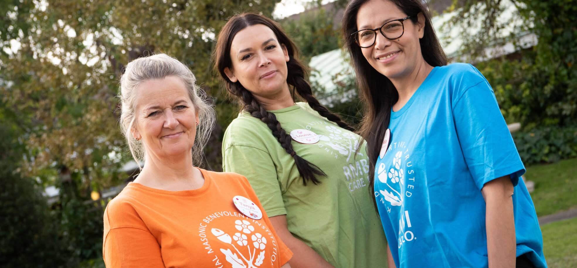 Three female staff members stand together, wearing bright orange, blue, and green shirts, smiling and enjoying each other's company at Prince George Duke of Kent Court care home.