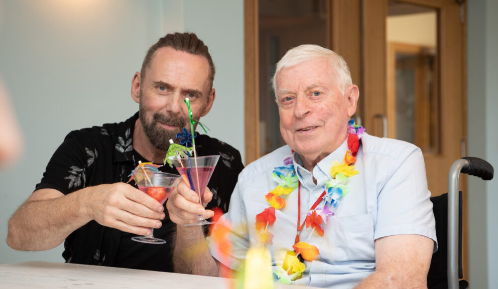 A man in a wheelchair takes a sip from his drink, exuding joy and contentment in a bright, cheerful environment with a member of staff.