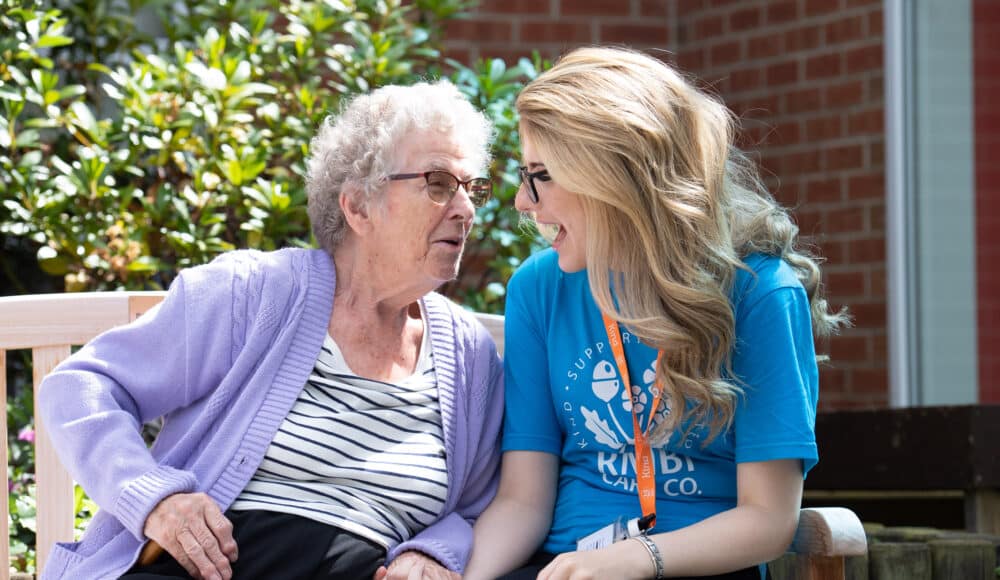 A young woman and an elderly woman sit side by side on a bench, enjoying each other's company in a serene outdoor setting.