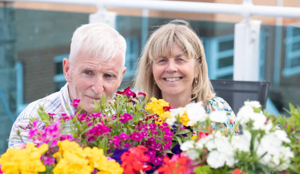 A couple smiles together, surrounded by vibrant flowers, capturing a joyful moment in their lives.