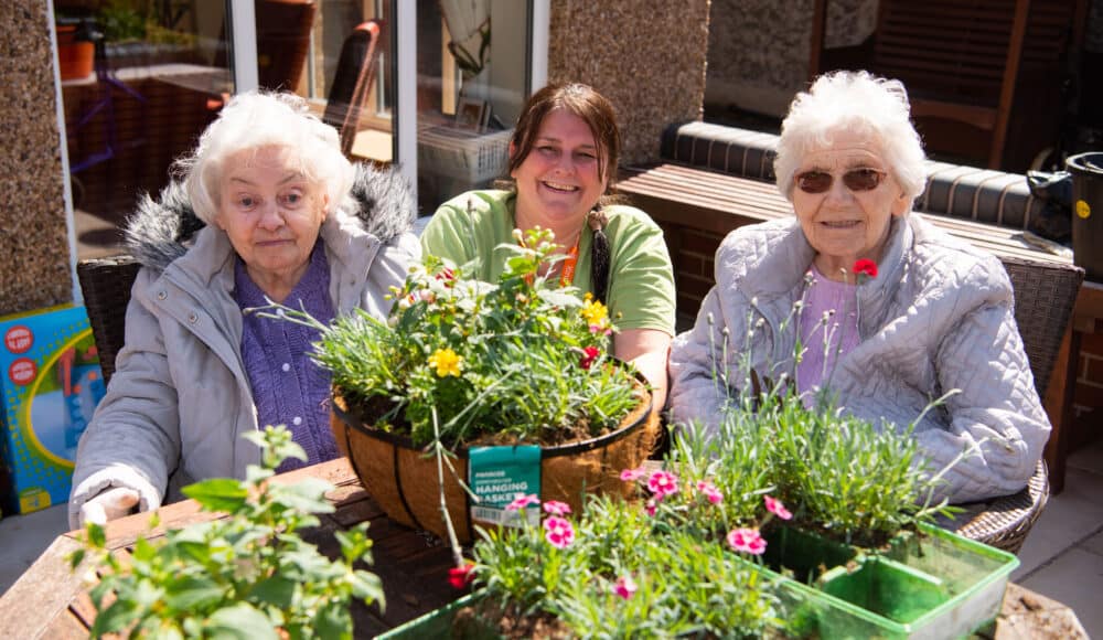 Two female residents and a female staff member sit around a table adorned with colorful flowers, sharing smiles and conversation.