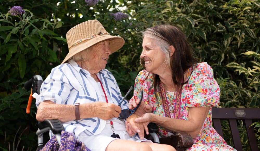 A female resident wearing a hat in a wheelchair besides a female staff memeber both smiling in a sunny outdoor setting.