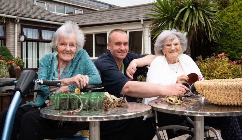 A male staff member and two female residents engaged in conversation while sitting at a table, sharing a moment together.