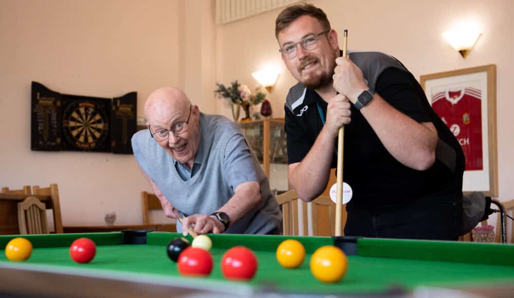 A male resident and a male staff member enjoying a game of pool at a table in a cozy room.