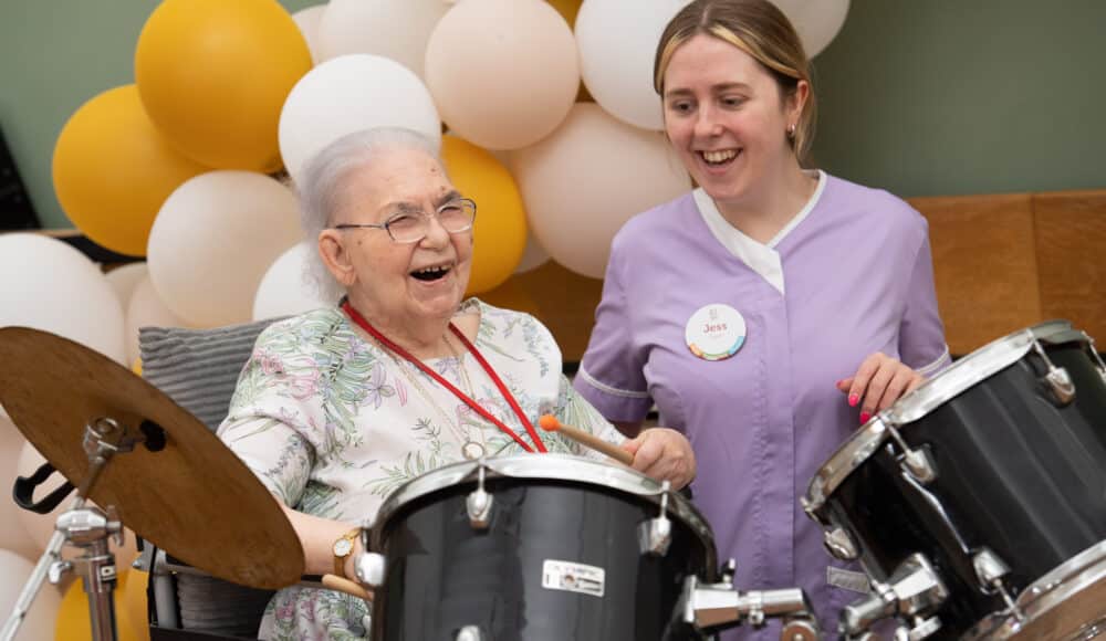 A smiling female resident plays the drums with a female staff member, both enjoying a lively moment of music and togetherness.
