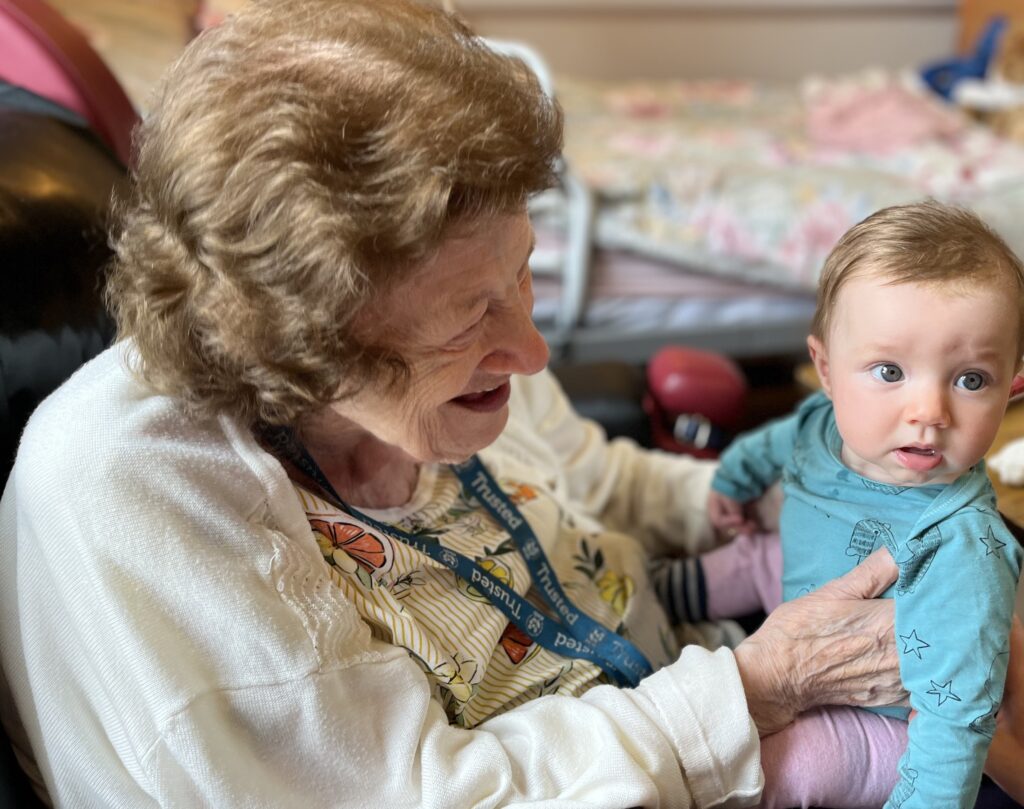 Resident Jean in a white top and a blue lanyard around her neck  at RMBI Home Prince George Duke of Kent Court, in Chislehurst is delighted to meet and hold baby Polly wearing a blue top.