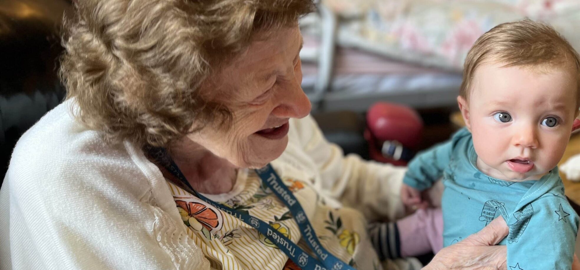 Resident Jean in a white top and a blue lanyard around her neck at RMBI Home Prince George Duke of Kent Court, in Chislehurst is delighted to meet and hold baby Polly wearing a blue top.