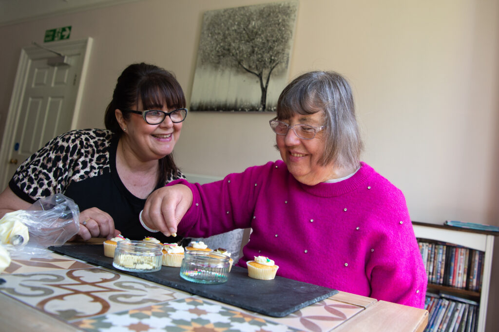 A staff member in a dark top with leopards printed sleeves and a resident in a pink jumper enjoy a delightful moment at a table adorned with colourful cupcakes, sharing smiles and laughter.