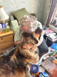 Resident Phyllis Mason, wearing glasses and sitting in her armchair, affectionately hugs Chewie. 