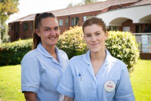 Two female staff members, dressed in light blue uniforms, smile at the camera in an outdoors location. There's a bush behind, and the care home.