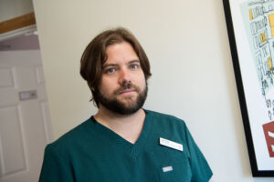 Jordi wears a nurse uniform and looks at the camera. He has long hair and a beard.