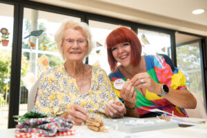 Resident Gladys, wearing a floral-patterned shirt and glasses. smiles next to Activities Coordinator Sharon, who has read hair and wears a colourful dress. They're knitting together. 