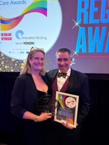 RMBI Care Co.’s Regional Manager for the central region and former Home Manager, Denise O’Brien, proudly holds up her trophy at the Great British Care Awards gala for the East of England, in Milton Keynes. Pictured here with her husband Neil O’Brien. They are attending the gala, and there is a big screen behind them. 