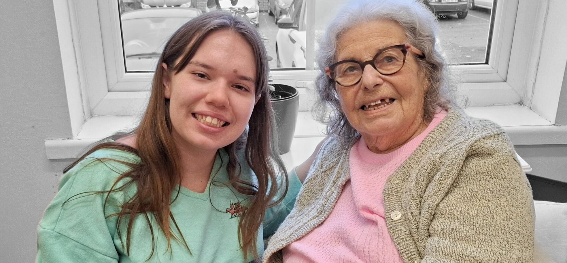 A young girl in a green jumper and her older pen pal in a pink yop and beige cardigan, and wearing glasses sit side by side, exchanging smiles and enjoying a heartfelt conversation.