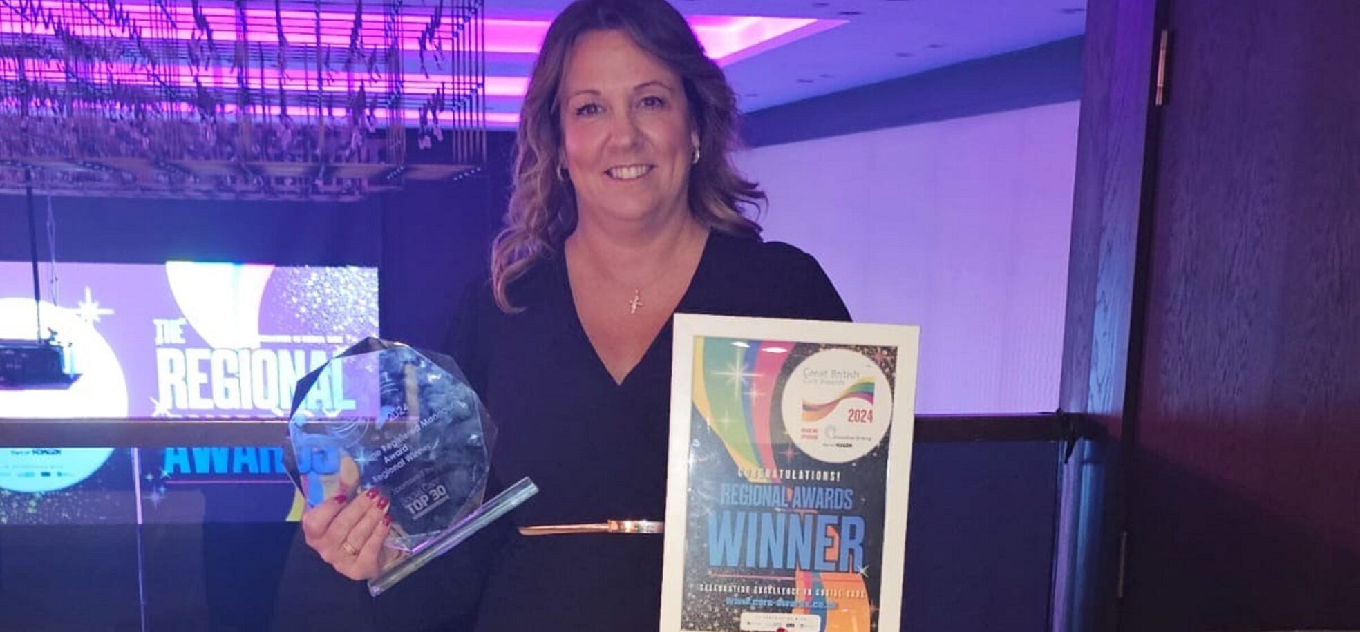 Nina Stephens, Prince George Duke of Kent Court's Home Manager in a blue pleated dress and gold belt holding her glass trophy and certificate, at the London Great British Care Awards with purple lighting.