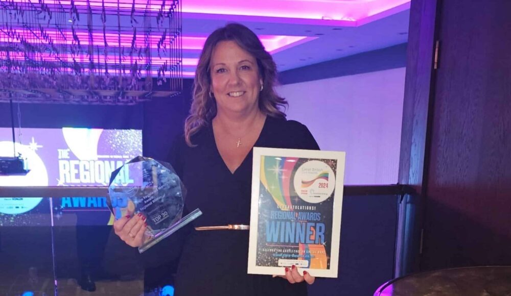Nina Stephens, Prince George Duke of Kent Court's Home Manager in a blue pleated dress and gold belt holding her glass trophy and certificate, at the London Great British Care Awards with purple lighting.