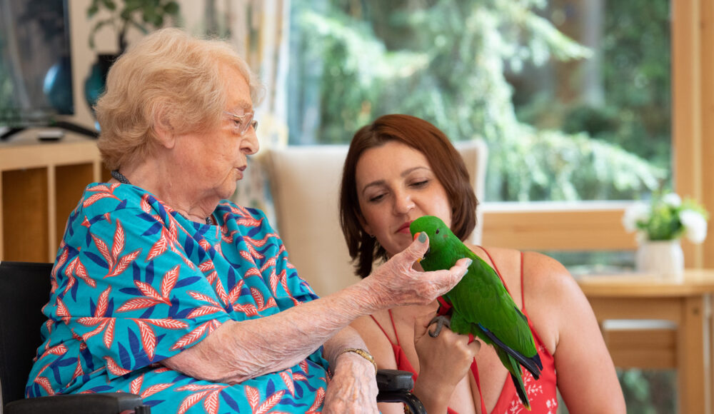 Resident Eunice feeds the green parrot with support from staff member Veronika.