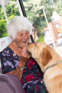 The resident, a short-haired lady wearing a dress with a floral pattern, smiles while holding the treat. Kobi is right next to her, looking at her eyes.