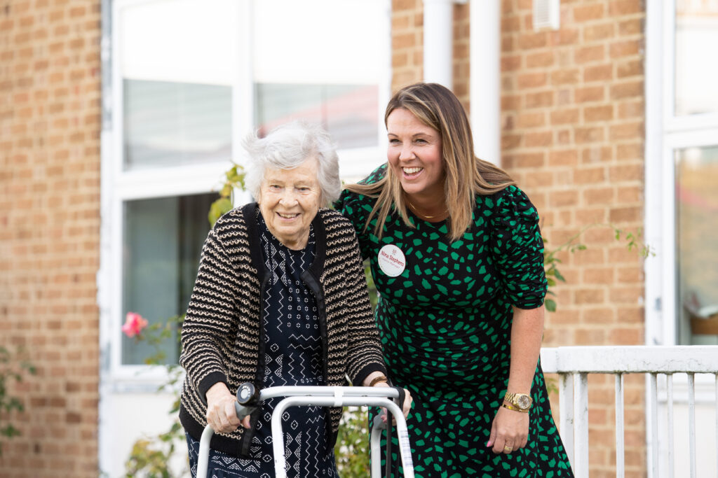 Nina Stephens, Prince George Duke of Kent Court's Home Manager in a green leopard dress with a female resident in a black and beige stripy cardigan with a walker, outside the care home.