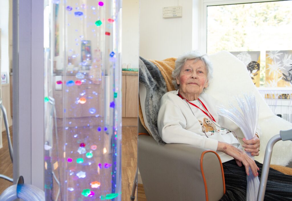 Resident Sheila in a white top sitting down and enjoying the colourful sensory light equipment in the dementia house at RMBI Care Co. Home Prince George Duke of Kent Court, in Chislehurst.