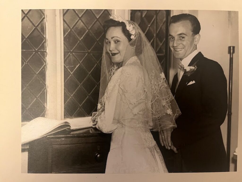 Black and white photo of  Pamela  in a white dress and headdress, and Tony in a suit, signing their name in a registry book  on their wedding day in 1955.