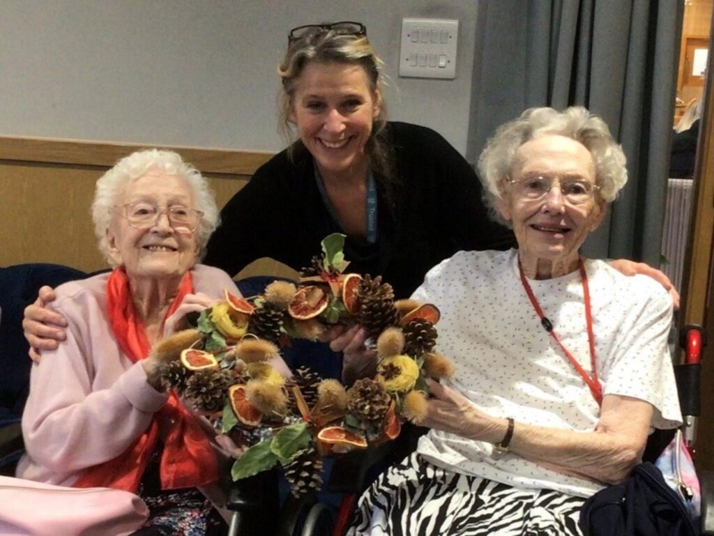 Two residents in wheelchairs and a staff joyfully hold a beautiful wreath, symbolizing friendship and unity in celebration.