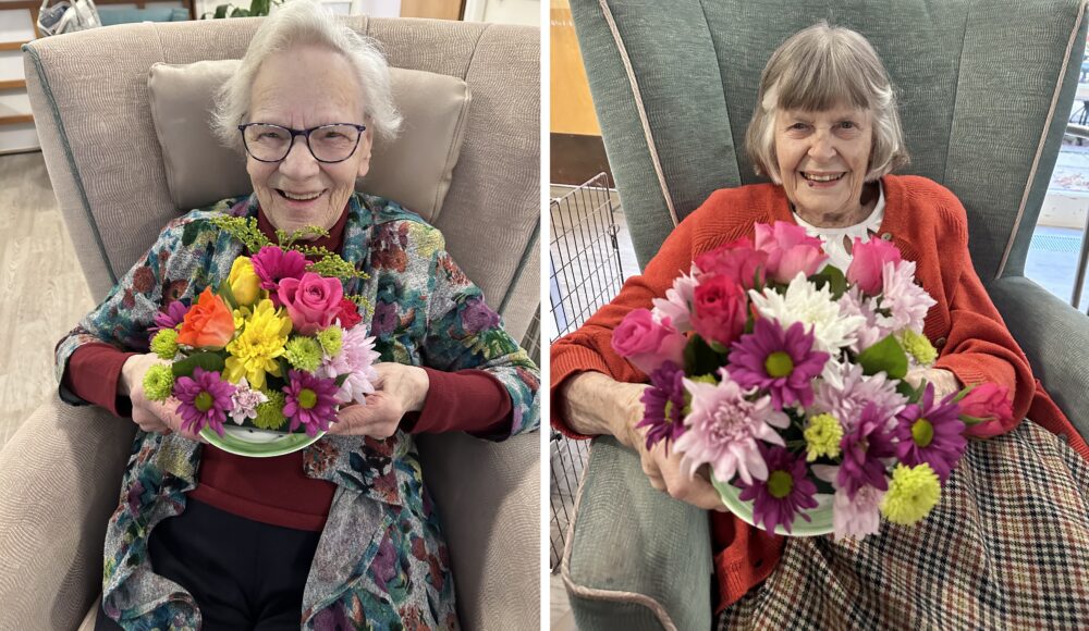 Two elderly women sitting in cozy armchairs, each holding a vibrant bouquet of flowers. The woman on the left wears glasses and a colorful shawl, smiling broadly while showcasing a mix of pink, yellow, and purple flowers in a green bowl. The woman on the right, dressed in a red sweater, also smiles while holding a bouquet filled with pink and white flowers, highlighting her joyful expression. The background features soft furnishings and warm lighting, creating a welcoming atmosphere.