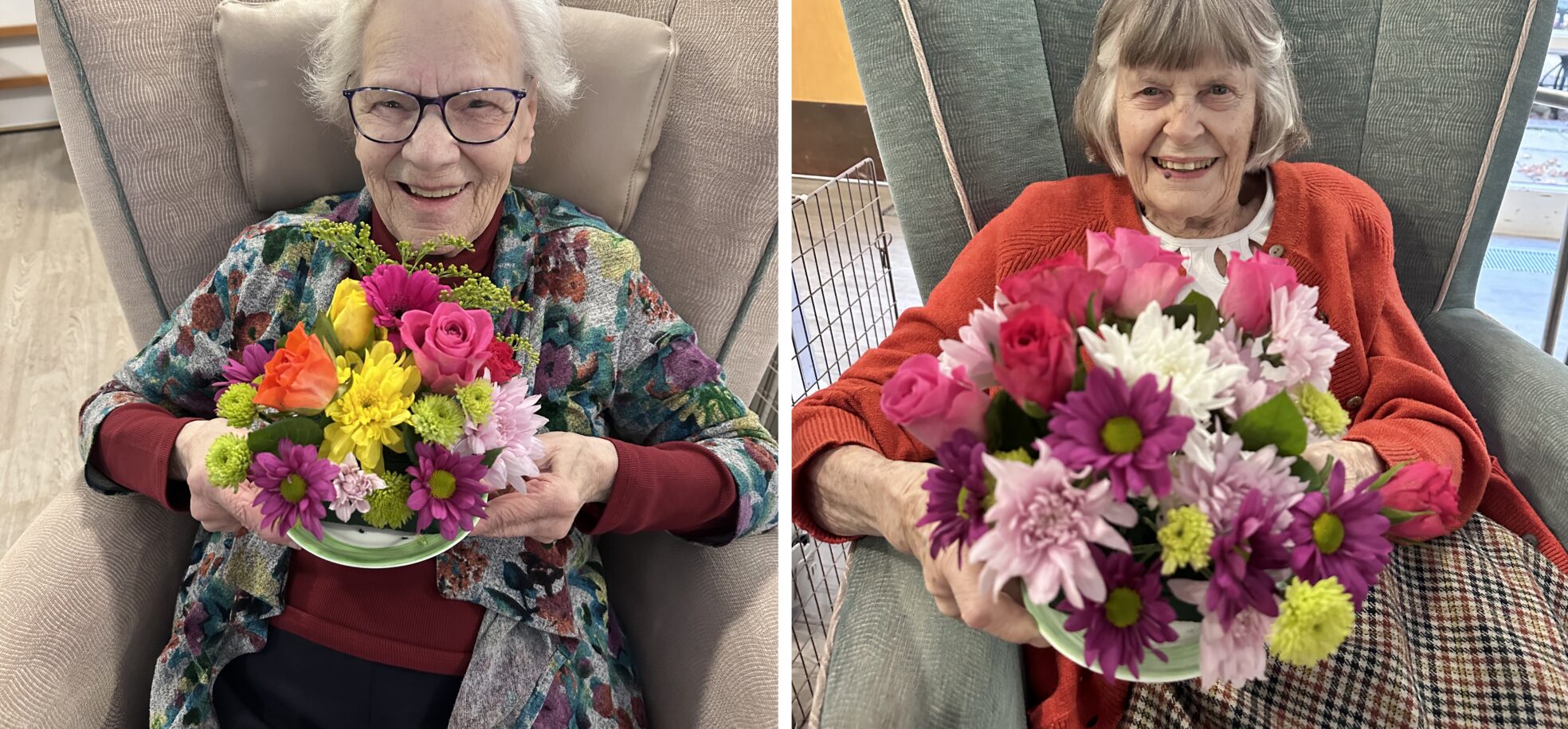 Two elderly women sitting in cozy armchairs, each holding a vibrant bouquet of flowers. The woman on the left wears glasses and a colorful shawl, smiling broadly while showcasing a mix of pink, yellow, and purple flowers in a green bowl. The woman on the right, dressed in a red sweater, also smiles while holding a bouquet filled with pink and white flowers, highlighting her joyful expression. The background features soft furnishings and warm lighting, creating a welcoming atmosphere.