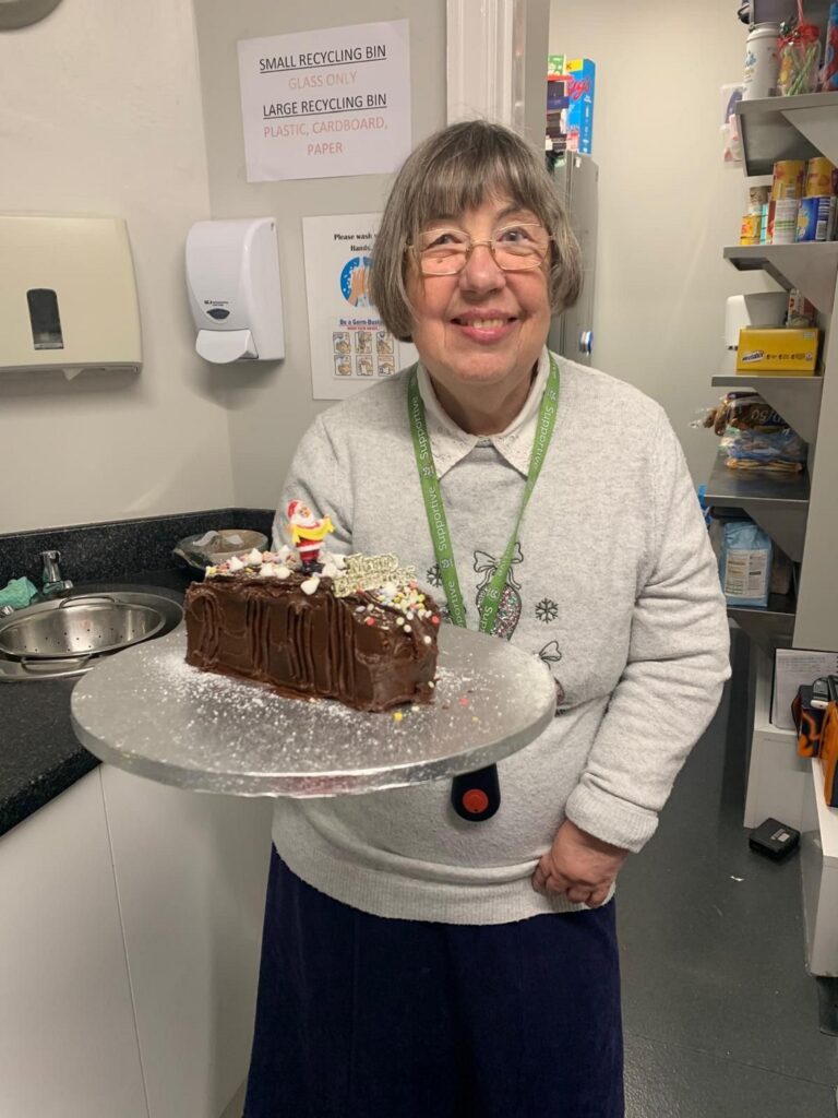 A resident smiles while holding a beautifully decorated cake in a cozy kitchen, surrounded by warm, inviting decor.