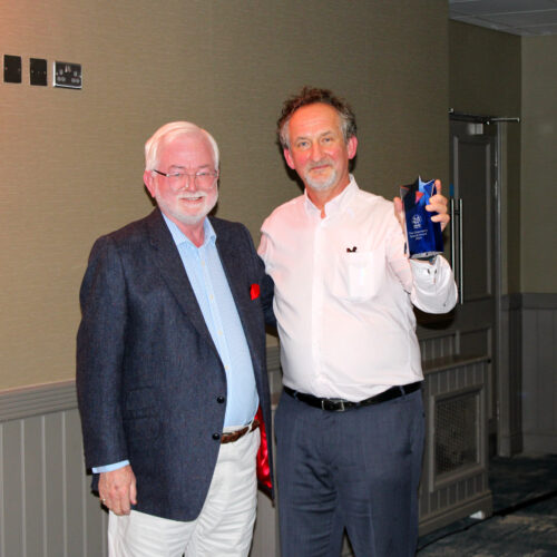 Two men are standing in a room, and one is presenting the other with a dark-blue award or prize. The man on the left, who seems to be receiving the award, is middle-aged, with short, graying hair and glasses. He is wearing a dark-navy or gray-blue suit jacket over a light patterned or striped shirt.  The jacket's lapels seem to match the color of the shirt. Light beige or cream-colored trousers complete his attire. His posture and expression suggest a pleasant reception of the award.

The man on the right, presenting the award, is also middle-aged with short brown hair. He is wearing a white, long-sleeved dress shirt and dark-colored pants, along with a dark belt. He is in the act of presenting a decorative, dark-blue trophy or a similar award, holding it somewhat prominently. His facial expression displays celebration and/or pride.

Both men are standing in a room with light beige or tan-colored walls.  The walls have a subtle vertical paneling effect with trim around the baseboard area.  There are small, square, light-reflective objects on the wall above the men, likely decor or signage.