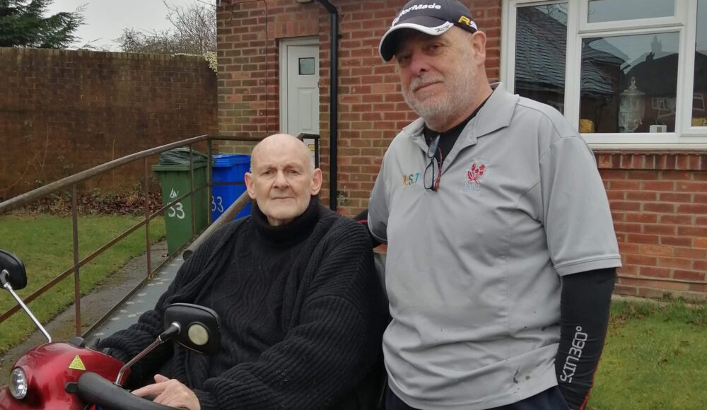 Staff member Philip in a blue cap, and grey polo shirt joyfully presenting Terry in a black coat while sitting down, a red motorized mobility scooter for better mobility.