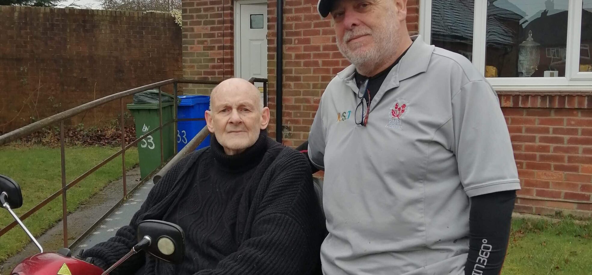 Staff member Philip in a blue cap, and grey polo shirt joyfully presenting Terry in a black coat while sitting down, a red motorized mobility scooter for better mobility.