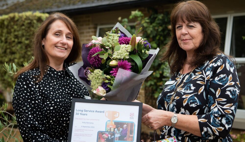 Training Manager Kelly Harrowing in a black dress with white dots celebrates her 30 years long service award with Home Manager Beverley Roberts in a dark floral dress at RMBI Care Co. Home Prince Michael of Kent Court, in Watford.