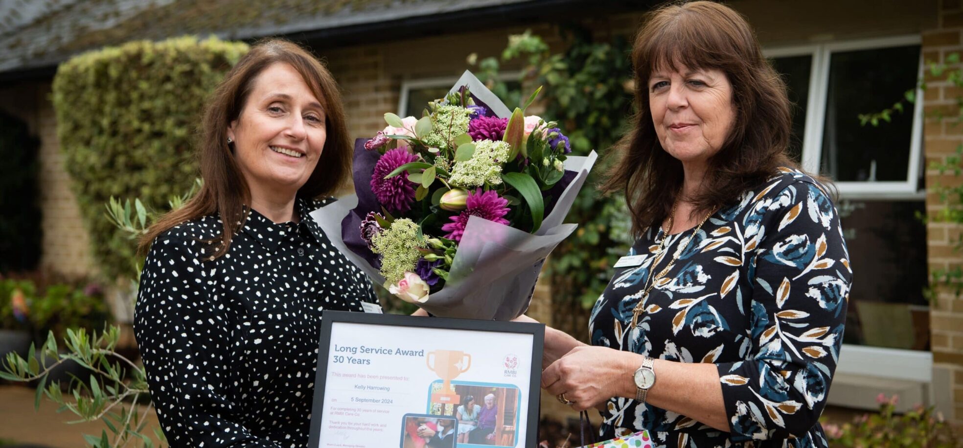 Training Manager Kelly Harrowing in a black dress with white dots celebrates her 30 years long service award with Home Manager Beverley Roberts in a dark floral dress at RMBI Care Co. Home Prince Michael of Kent Court, in Watford.