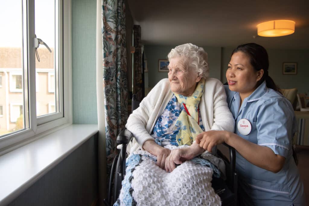 In a wheelchair, an older lady looks outside the window. Next to her, with a knee on the floor, is a staff member wearing her uniform. The lady is white and the staff member is Asian.