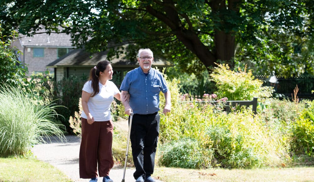 Staff member Kumare in a white t shirt and borwn trousers, and resident Graham in a blue shirt and black trousers enjoy a relaxing afternoon walk through the gardens at RMBI Care Co. Home Connaught Court, in York