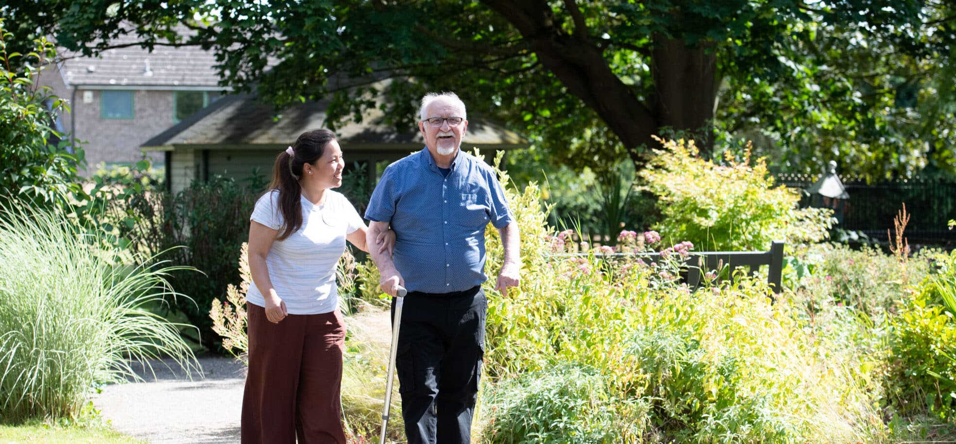 Staff member Kumare in a white t shirt and borwn trousers, and resident Graham in a blue shirt and black trousers enjoy a relaxing afternoon walk through the gardens at RMBI Care Co. Home Connaught Court, in York