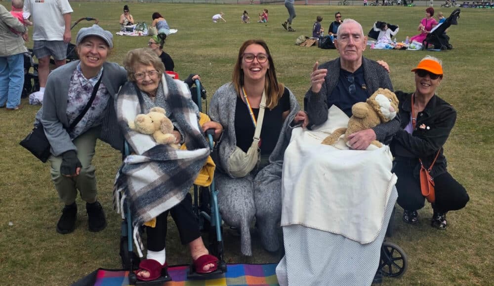 Two residents sitting, one activities coordinator and two volunteers crouching at the teddy bears' picnic. There are other people enjoying their picnics in the background.