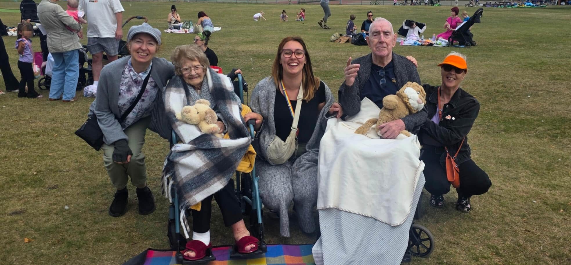 Two residents sitting, one activities coordinator and two volunteers crouching at the teddy bears' picnic. There are other people enjoying their picnics in the background.