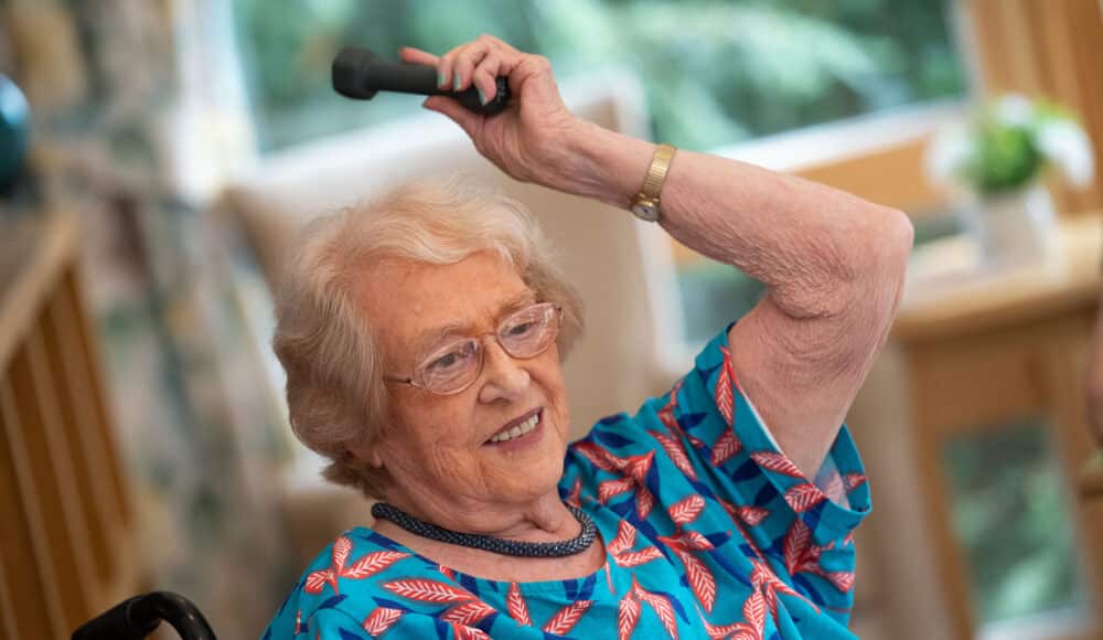 Resident in a bright orange and blue patterned blouse raises her arm whilst holding a light hand weight at James Terry Court care home.