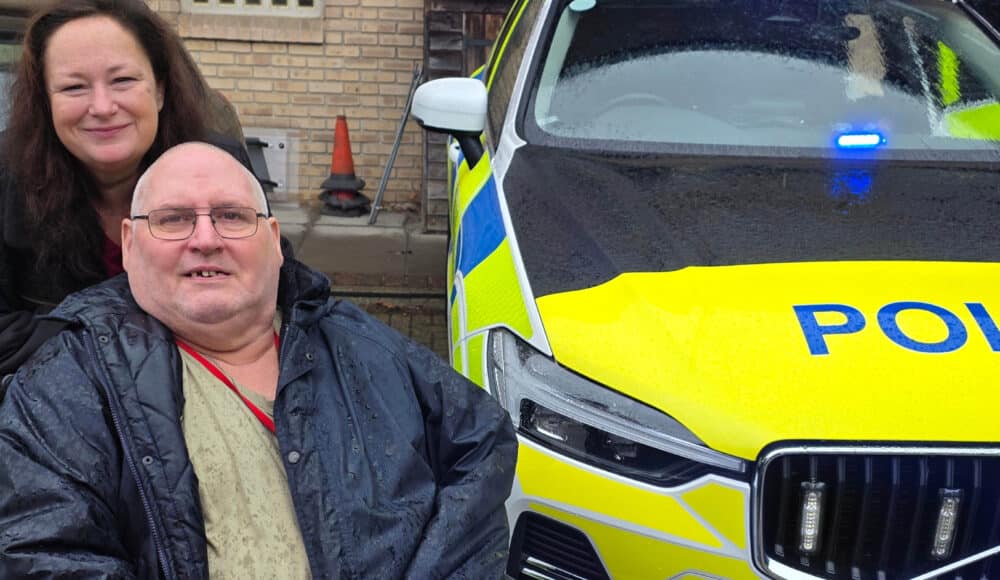 A male resident sitting in a wheelchair and smiling next to a police car. A female staff member is standing behind him, also smiling.