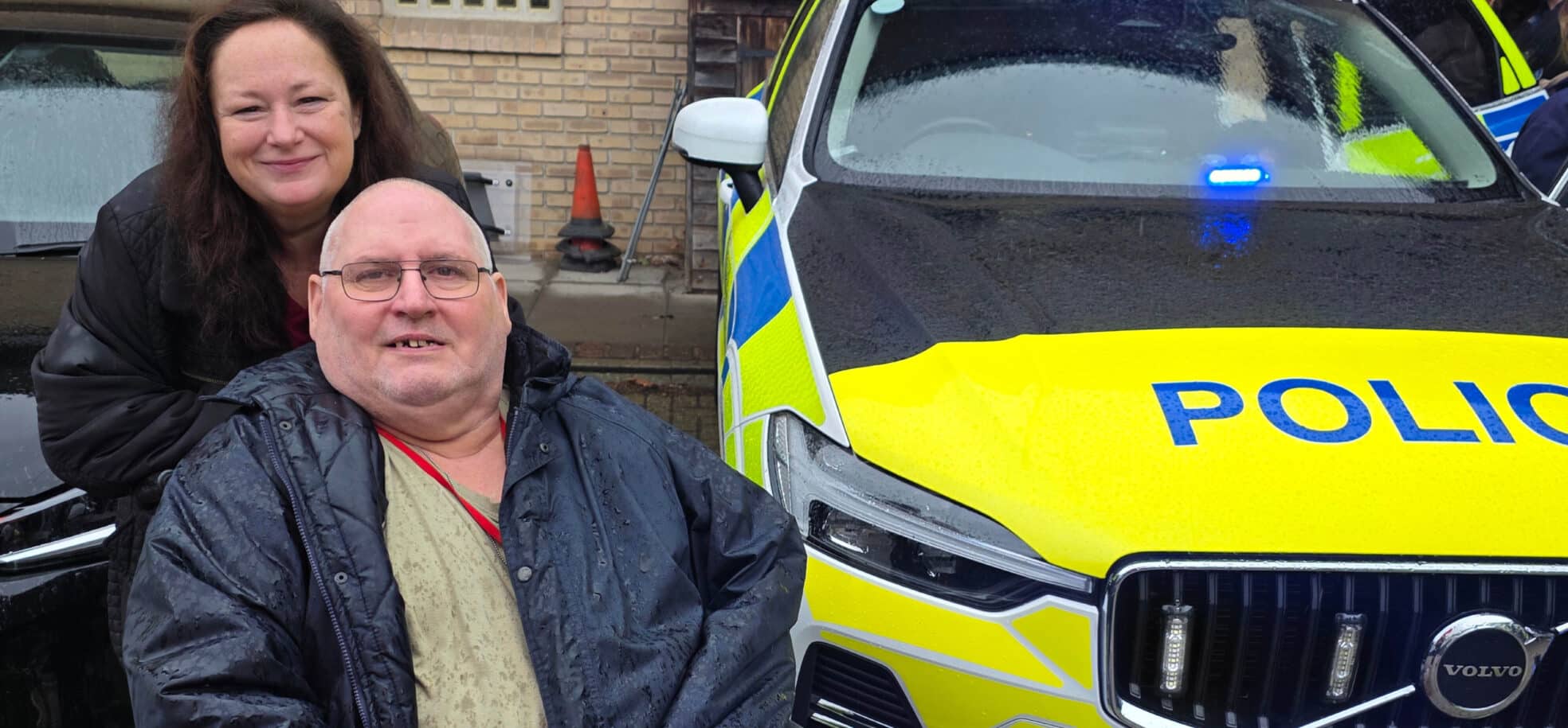 A male resident sitting in a wheelchair and smiling next to a police car. A female staff member is standing behind him, also smiling.