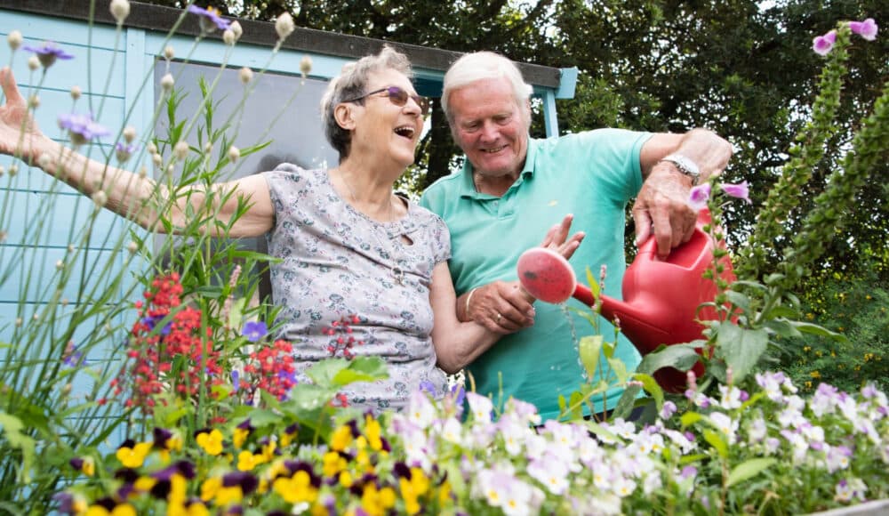 A female and a male resident smile while watering one of the flower beds of their care home. They are in Hove, England.