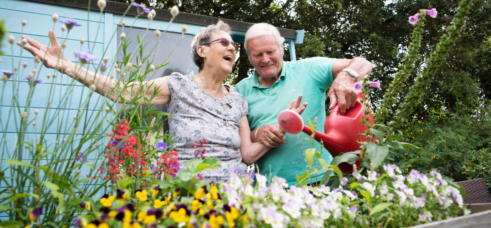 A female and a male resident smile while watering one of the flower beds of their care home. They are in Hove, England.