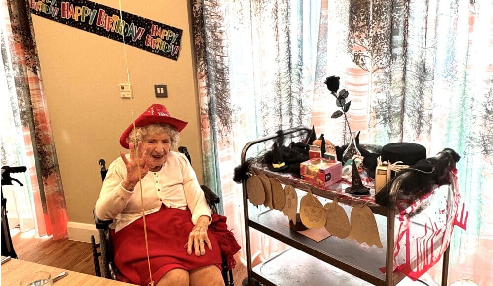 Older woman in red dress and hat waves cheerfully, seated by a table in a care home. A festive "Happy Birthday" banner hangs above. Decorated cart nearby.